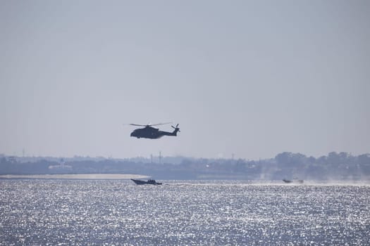 Military helicopter flying over the ocean, with patrol boats assisting in surveillance and potential rescue operations