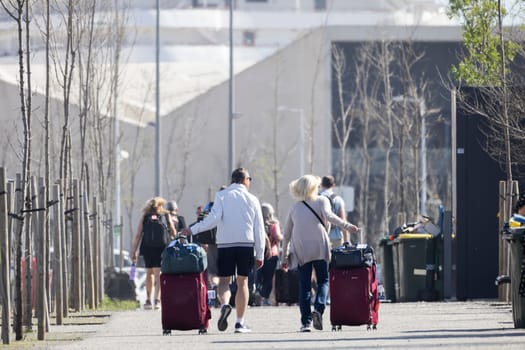 Tourists pulling rolling suitcases and carrying travel bags are walking outside an airport terminal on a sunny day