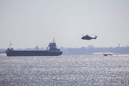 Military helicopter patrolling over cargo ship and motorboat sailing on a river