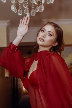 Model in red dress, elegant young woman standing near exquisite crystal chandelier. Beautiful girl with high hairstyle, shooting in hotel. Model with evening make-up and hairstyle, looking at camera