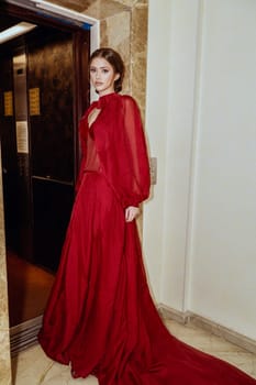 Model in red dress, elegant young woman with evening make-up and hairstyle. girl stands near open elevator, looks at camera. Beautiful girl in long evening dress, shooting in hotel interior.