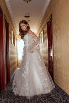 A young bride in a long classic dress with open alechi stands in the hotel corridor. She is in a silk dress with lace. She has long hair styled in a wave.