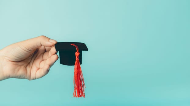 Female hand holding graduation cap on blue background. Symbol of education, achievement, academic success, student life, and graduation ceremony.