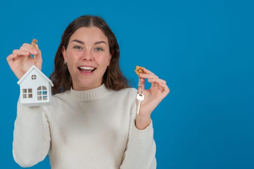 Happy young woman holding a house model and keys, expressing excitement. She wears a white sweater and stands against a blue background. Concept of home ownership, real estate, or investment. Copy space