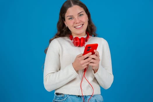 Young woman wearing red headphones around her neck, smiling while looking at her smartphone. She appears happy and excited,possibly reacting to a message or content.looking at camera, Blue background.