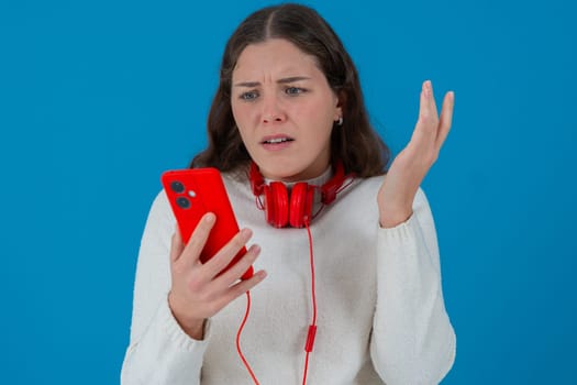 Caucasian woman looks shocked while reading bad news on her smartphone. She wears headphones and a white sweater, reacting with concern against a solid blue background.