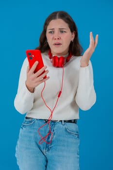 Caucasian woman looks shocked while reading bad news on her smartphone. She wears headphones and a white sweater, reacting with concern against a solid blue background.Vertical