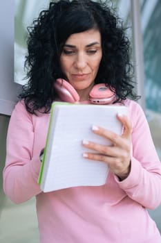 Caucasian sporty woman in a pink hoodie writing in a notebook outdoors. Wearing headphones around her neck, she appears focused and relaxed after a workout in an urban setting. Vertical