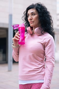 Caucasian athletic woman wearing a pink hoodie and headphones around her neck holds a pink water bottle. She stands in an urban setting, appearing thoughtful and focused after a workout. Vertical