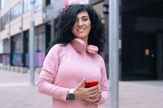 Individual portrait of a Caucasian woman in a pink hoodie smiling at the camera. She holds a red smartphone and wears headphones around her neck in a casual urban outdoor setting.