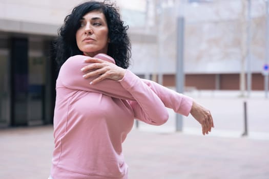 Caucasian adult woman with curly black hair stretching her arms outdoors in an urban setting.she is wearing a pink sweatshirt and exercising for a healthy lifestyle, fitness, and wellness in the city