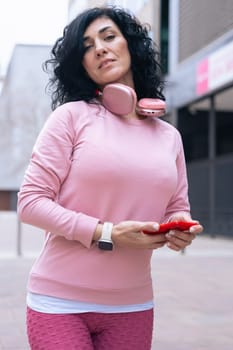 Individual portrait of a Caucasian woman in a pink hoodie smiling at the camera. She holds a red smartphone and wears headphones around her neck in a casual urban outdoor setting.Vertical