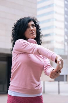 Caucasian adult woman with curly black hair stretching her arms outdoors in an urban setting.she is wearing a pink sweatshirt and exercising for a healthy lifestyle, fitness, and wellness in the city