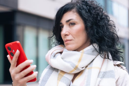 Serious caucasian woman standing outdoors in urban setting, wearing coat and scarf, using her red smartphone. She is serious and relaxed, enjoying a casual moment on a cold day in the city.