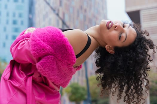 Graceful dancer with curly hair wearing a pink coat, expressing movement and energy in an urban environment. Modern fashion, contemporary dance, and artistic performance against a city backdrop.