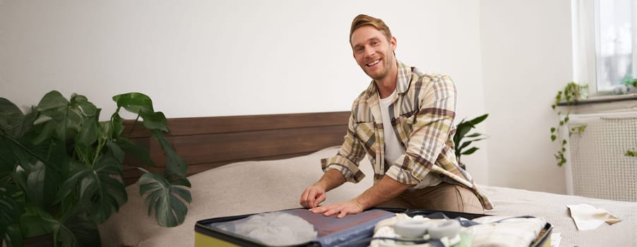 Portrait of young handsome bearded guy, tourist going on holiday, fastening his clothes in suitcase, packing for vacation, going on business trip, sitting in bedroom.