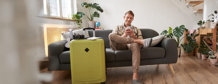 Handsome young man sits on sofa with suitcase, holds smartphone, books hostel or holiday vacation, chats on mobile phone. Tourism and travelling concept