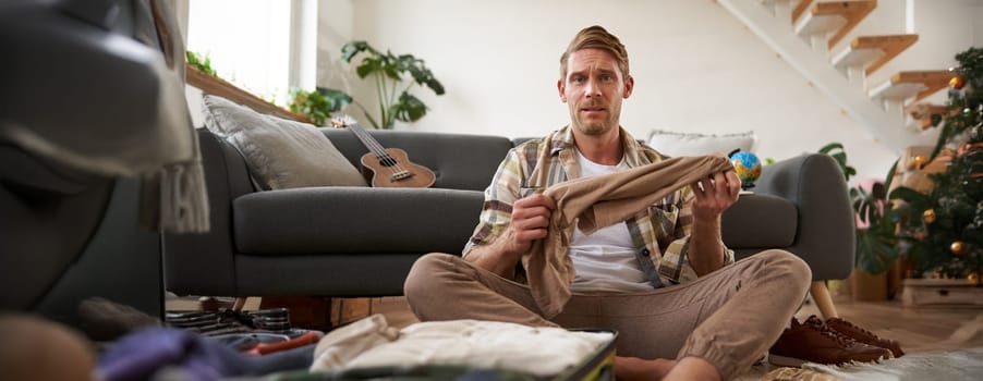 Portrait of confused man, sitting in living room with suitcase, holding pair of trousers, looking clueless, struggling to pack for holiday. Tourism and travelling concept