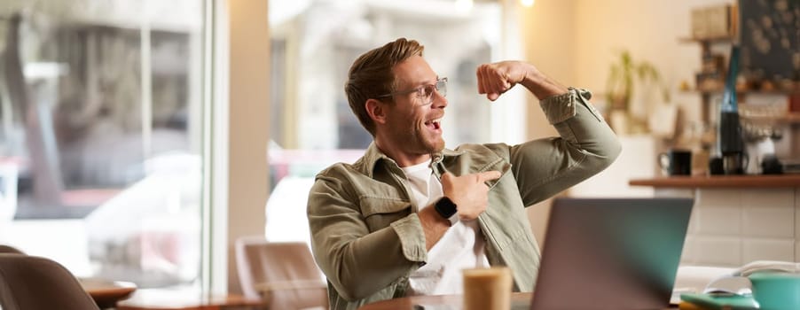 Portrait of young and healthy masculine man, shows his biceps to someone over video chat, using laptop for a call, pointing at his muscles and smiling pleased, sitting in cafe.