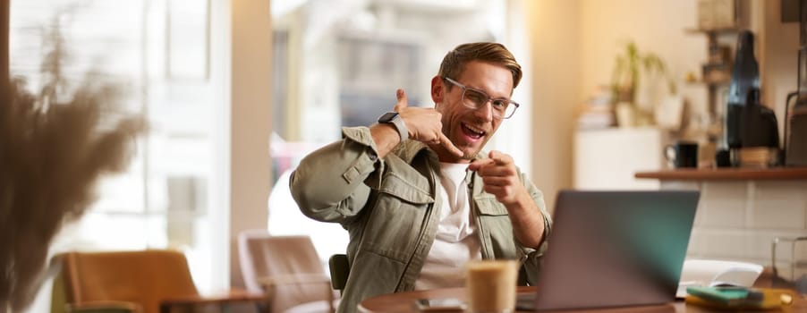Portrait of cheerful, flirty guy in glasses, sitting with laptop computer in cafe, showing phone call hand gesture and winking at you, giving his number.