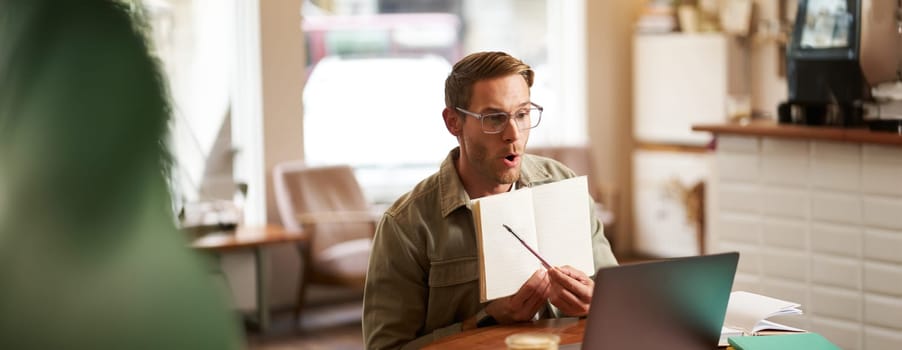 Portrait of handsome young man giving lessons online, showing exercises in his notebook, looking at laptop, recording video, sitting in cafe, quite co-working space.