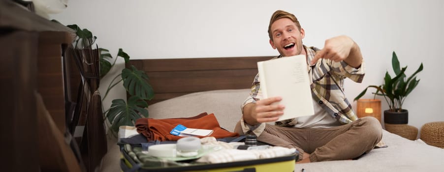 Portrait of young man pointing at his list of items, packing clothes on vacation, showing notebook, sitting near opened suitcase. Lifestyle and people concept