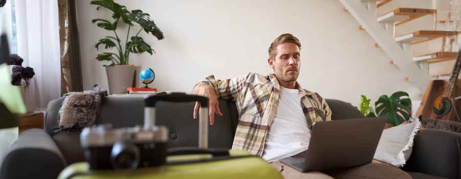 Happy man booking holiday online on laptop, paying for vacation on travel agency website, buying flight tickets in internet, sitting in living room with suitcase.