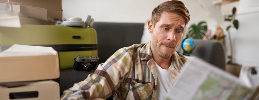 Portrait of man looking confused at travel map, choosing route, going on holiday, preparing for vacation, sitting in living room with suitcase.