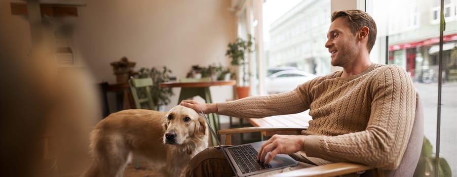 Portrait of young man, digital nomad, sitting in cafe near window, using laptop, working with his pet, petting golden retriever while sitting in chair with computer on laps.