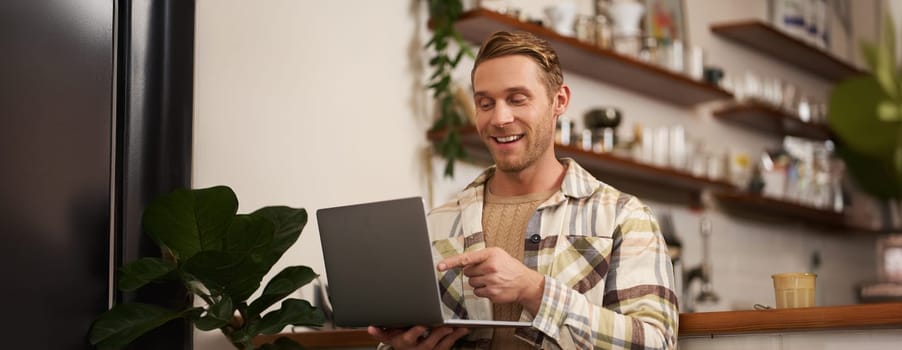 Image of young man in coffee shop, connects to video call, talking online, chats with friends via laptop and smiles.