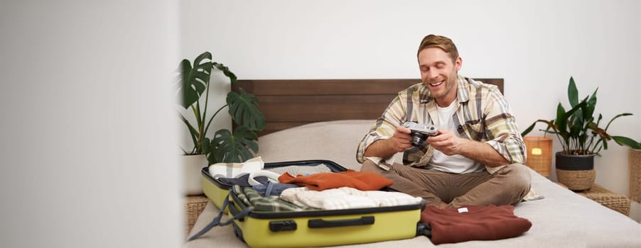 Portrait of happy, smiling young man sitting on bed, unpacking his suitcase after a holiday, tourist looking at his photo album on camera.