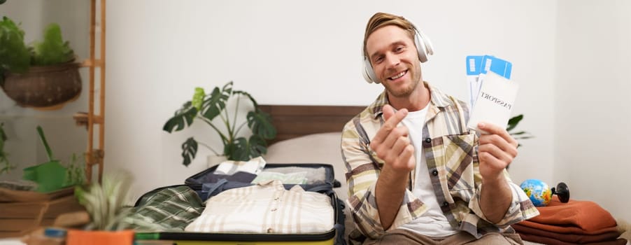 Image of handsome young man, tourist shows two plane tickets and passport, travels on holiday, goes on vacation, packs suitcases.