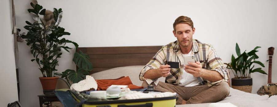 Portrait of happy man in suitcase, transferring money, buying tickets online, sitting on bed with credit card and smartphone app, paying online.