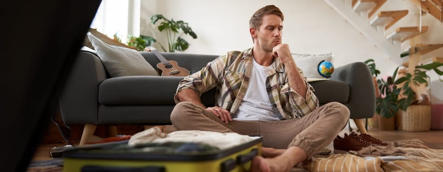 Portrait of stressed young man packing his suitcase, deciding what clothes bring on vacation, thinking while looking at luggage. Tourism concept
