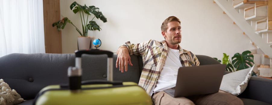 Portrait of happy young man, tourist booking his holiday with travel agency, looking at laptop, packed suitcase to go on vacation.