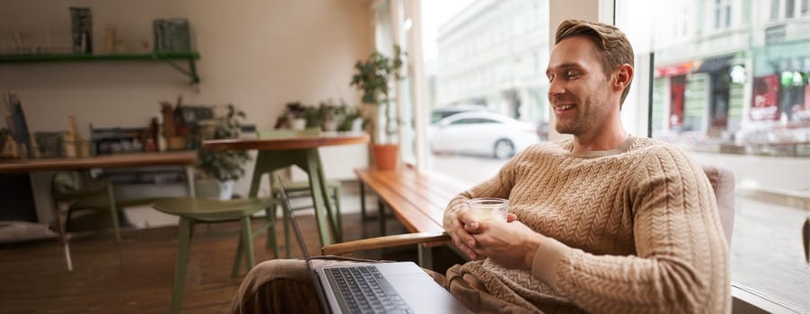 Portrait of handsome young man sits in cafe, drinks coffee and watches video on laptop, looking at screen with happy smile, relaxing in co-working space.