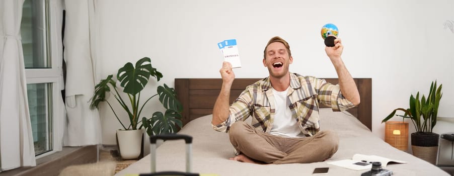 Portrait of cheerful, happy tourist, man sitting with plane tickets and a globe, packed suitcase for travelling around the world, going on vacation, excited about his holiday.
