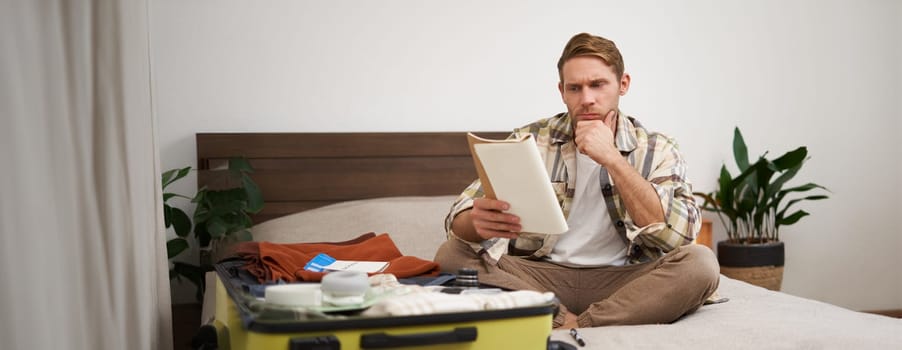 Portrait of man with suitcase, sits in room and looking serious at his notes, packing for holiday, checking list of items in notebook. Copy space