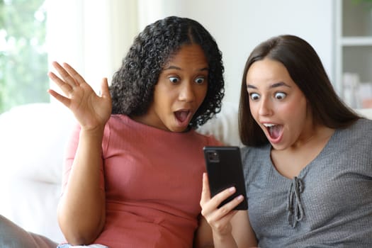 Amazed interracial friends checking cell phone content sitting on a couch at home