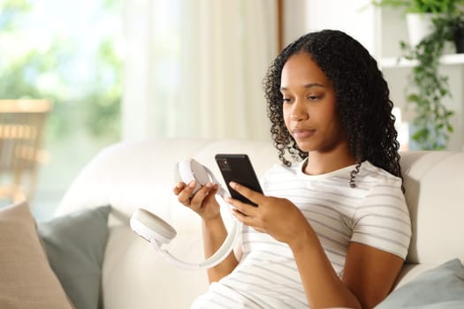 Black woman pairing wireless headphone with smart phone sitting on a couch at home
