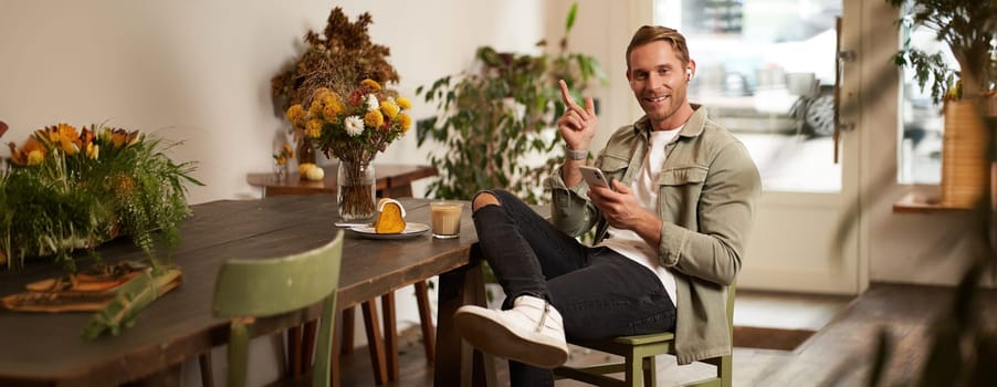 Portrait of happy handsome young man, sitting in cafe with mobile phone, wearing wireless headphones, listening to music or podcast while relaxing in coffee shop.
