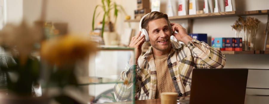 Lifestyle portrait of happy, excited young man, sitting in coffee shop with laptop and headphones, dancing on his chair and enjoying great quality sound, streaming favourite songs.