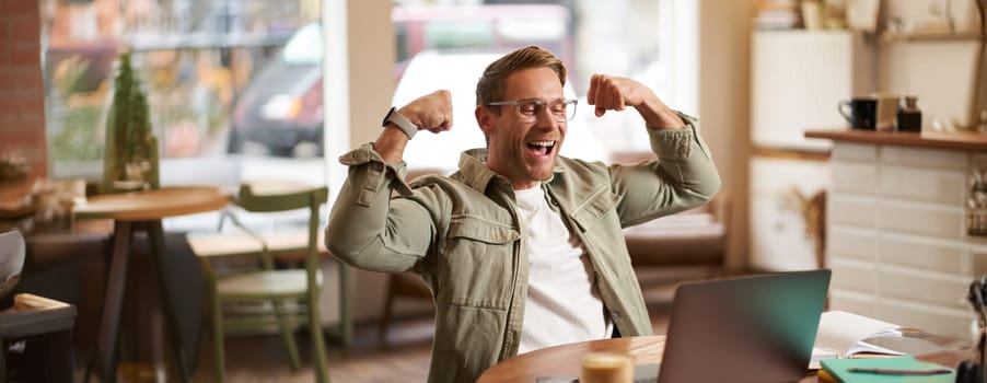 Portrait of happy young man in glasses rejoicing, sitting in cafe, watching sports game on laptop, celebrating, showing big muscles and laughing.