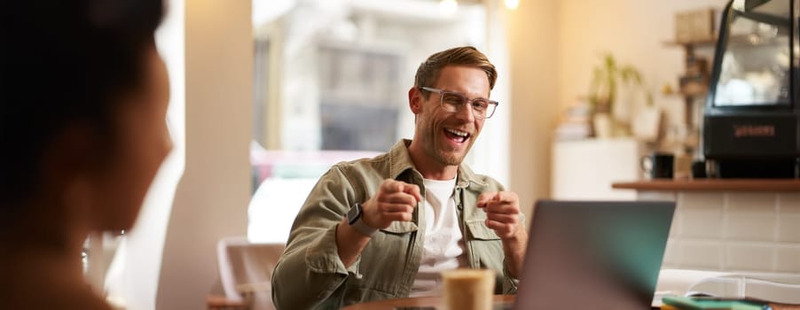 Happy young man in glasses, freelancer sitting in cafe with laptop, drinking coffee, pointing fingers at camera and smiling cheerfully.