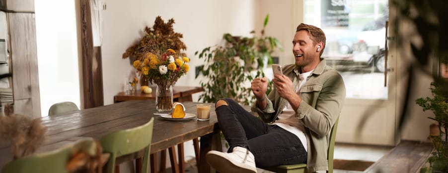 Portrait of happy handsome young man, sitting in cafe with mobile phone, wearing wireless headphones, listening to music or podcast while relaxing in coffee shop.