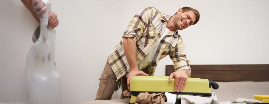 Image of young man, tourist struggling to close his suitcase full of clothes, pushing on bag, trying to press items inside luggage to shut the zipper, packing for travelling abroad, going on holiday,