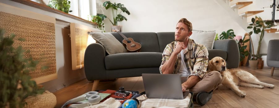 Portrait of young man, traveller going on holiday with a dog, packing his suitcase, sitting with laptop on his luggage, thinking, making online booking.