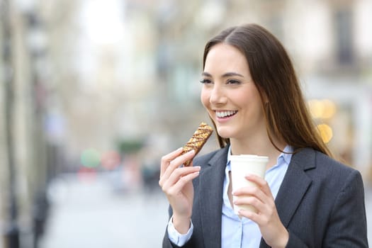 Happy executive in the street eating cereal bar and drinking coffee walking