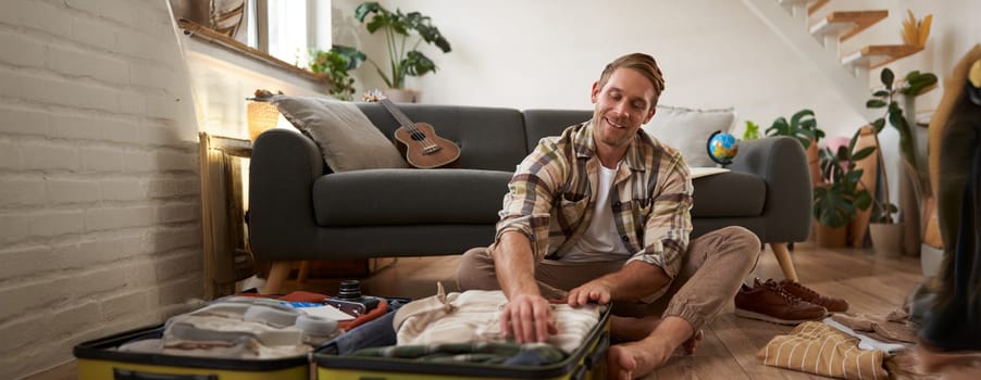 Image of young man, traveller, sitting on floor with opened suitcase, packing clothes for a trip, going on holiday. Tourism concept