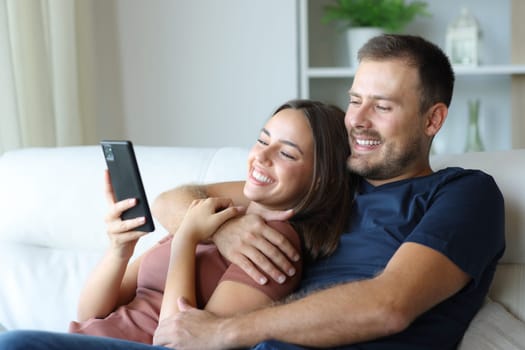 Happy couple using smart phone hugging sitting on a sofa at home